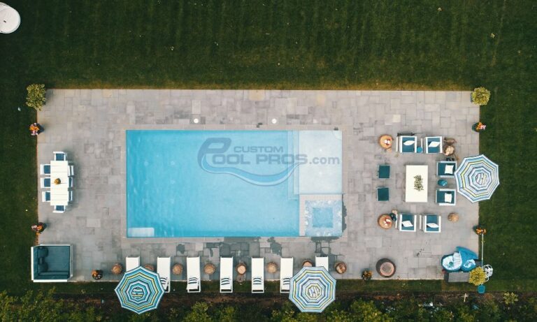 Aerial view of a modern backyard pool area featuring a rectangular swimming pool, surrounded by stone patio, lounge chairs, and colorful umbrellas. The space includes a dining area with a table and chairs, complemented by potted plants for a refreshing outdoor ambiance, inground pool builders