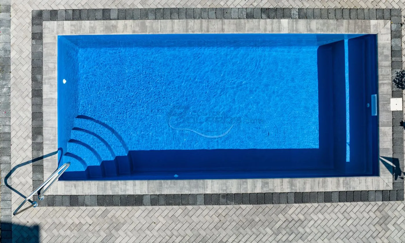 Blue swimming pool with a textured surface, surrounded by a gray stone deck, featuring curved wave patterns along one side, luxury pool builder.