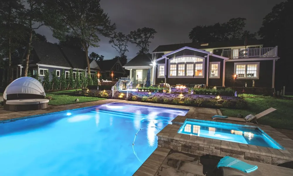 Luxurious backyard at night featuring a sparkling blue swimming pool, a spa, and a beautifully lit modern home surrounded by lush landscaping, Swimming pool companies in New Jersey