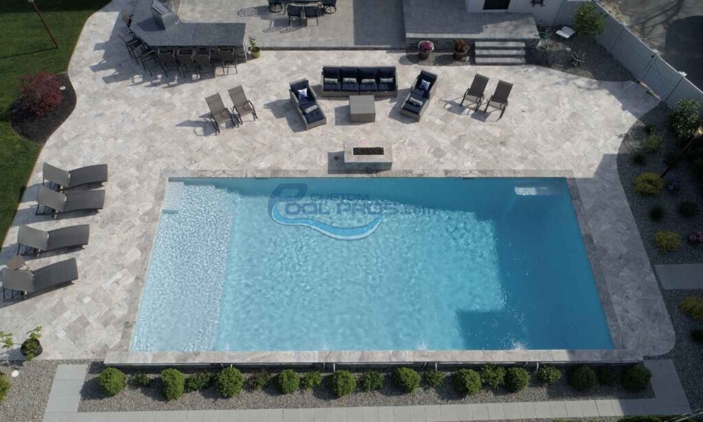 Aerial view of a modern outdoor swimming pool surrounded by lounge chairs and a seating area, featuring stone decking and neatly trimmed hedges. Ideal for relaxation and leisure activities, pool builder, inground swimming pool