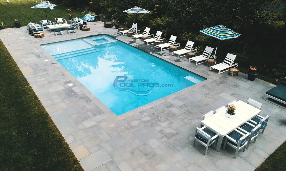 Aerial view of a modern swimming pool surrounded by lounge chairs and umbrellas, set in a landscaped outdoor area. The pool features a built-in spa and is ideal for relaxation and leisure activities, pool contractor