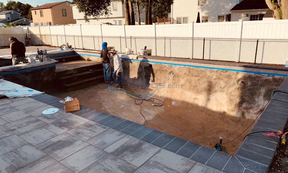 Workers excavating a swimming pool site, with tools and equipment visible, surrounded by a partially constructed patio and fencing, luxury pool builder